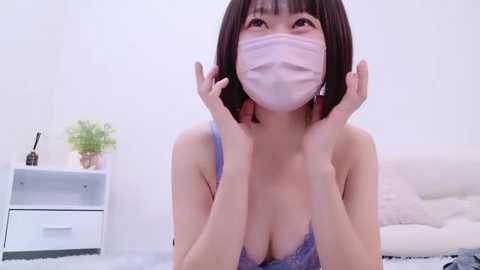 Media: Video of an Asian woman with short dark hair, wearing a face mask, a blue lace bra, and a white top, kneeling on a white bed, holding her face. Background includes a white dresser with a plant and a bottle.