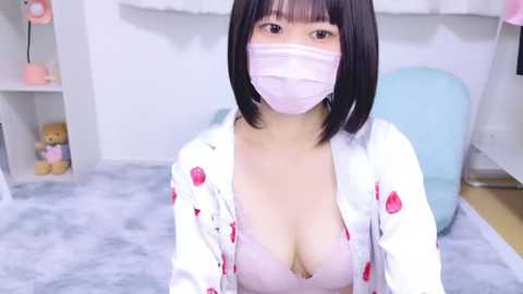 Media: Video of an East Asian woman with short black hair, wearing a pink mask, a white robe with red heart patches, and a pink bra, seated on a light blue chair in a room with a white wall and a gray carpet.