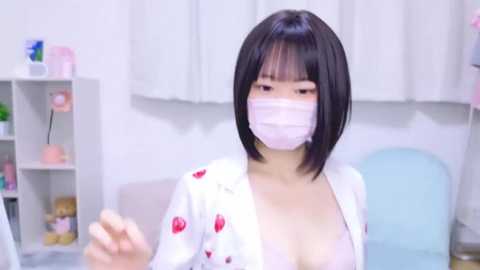 Media: Video of an East Asian woman with straight black bob hair, wearing a white robe with red cherries, pink face mask, and light pink bra. Background includes a white wall, shelves with pink objects, and a light blue chair.