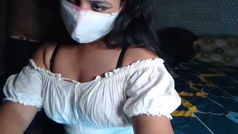 Media: A video of a woman with medium skin tone, wearing a white off-shoulder blouse, white mask, and black bra straps, lying on a bed with a geometric-patterned blue and yellow quilt.