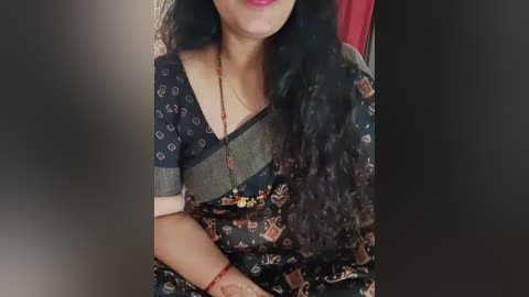 Media: Video of a South Asian woman with medium skin tone, wearing a dark, patterned sari with a gold necklace, seated indoors with a red curtain in the background.