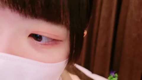 Media: Video of a young Asian woman with short, straight black hair and a white surgical mask covering her mouth, looking tired with half-closed eyes. Background features a wooden door and a purple flower.