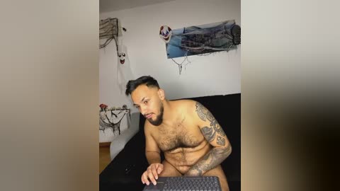 Media: Video of a muscular, tattooed, bearded Latino man with short black hair, shirtless, sitting on a black bed in a minimalistic, white-walled room, holding a black laptop.