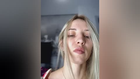 Media: Video of a young woman with straight, platinum blonde hair, wearing a red top, puckering her lips in a playful manner. Background is blurred with muted colors.