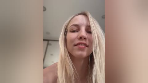 Media: Video of a young, fair-skinned, blonde woman with straight hair, smiling, standing in a dimly lit, modern room with white walls and a white wardrobe in the background.