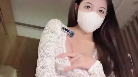 Media: A video of a young East Asian woman with long, dark hair, wearing a white lace top, white face mask, and holding a white device. She is indoors, with beige curtains and wooden floors in the background.