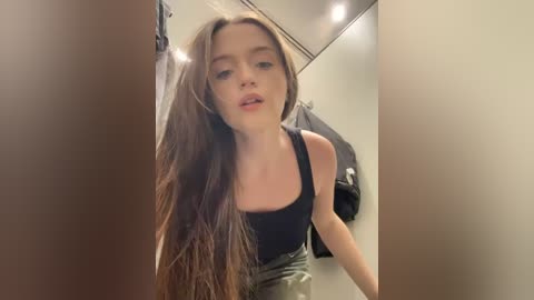 Media: Video of a young Caucasian woman with long brown hair in a black tank top, leaning forward in a dimly lit, beige-tiled bathroom.