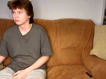 Media: Video of a young Caucasian man with tousled brown hair, wearing a grey t-shirt and striped pajama pants, sitting on a brown suede couch, looking contemplative.