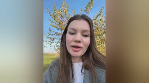 Media: Video of a young Caucasian woman with long brown hair, light skin, and a neutral expression, wearing a gray jacket over a white shirt. She stands in a field with a tree in the background under a clear blue sky.