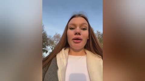 Media: Video of a fair-skinned woman with straight brown hair, wearing a grey jacket over a white top, against a snowy outdoor background.