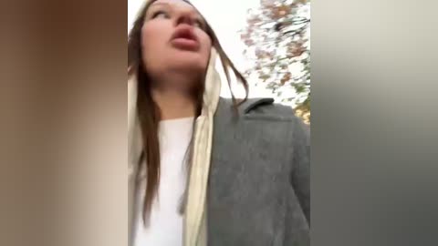 Media: A video of a young woman with straight brown hair, fair skin, and light makeup, wearing a white top and gray jacket, looking upward. Background shows blurred autumn trees.