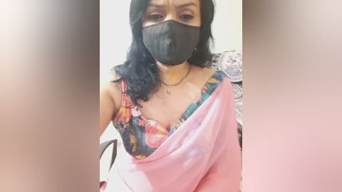 Media: Video of a woman with light skin, dark hair, and a black face mask, wearing a pink sari with a colorful blouse, in an indoor setting.