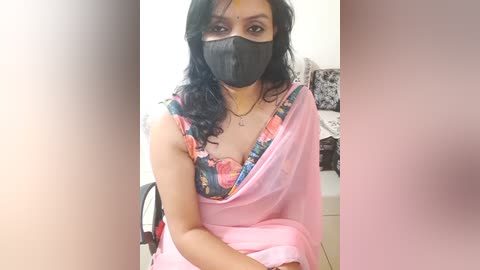 Media: A video of a South Asian woman with medium skin tone, wearing a pink saree, floral bra, black mask, and black hair, sitting indoors, blurred background.