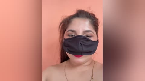 Media: Video of a woman with medium skin tone and straight dark hair, wearing a black face mask and red lipstick, against a peach-colored background.