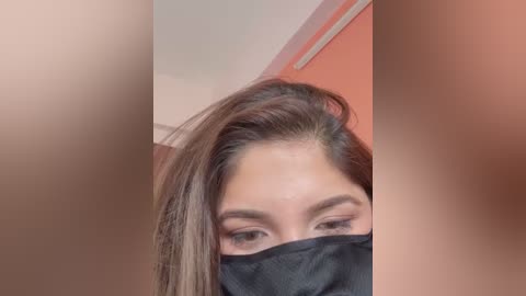 Media: A video of a woman with long brown hair, wearing a black face mask, her eyes partially visible, in a blurred, orange-lit bathroom setting.