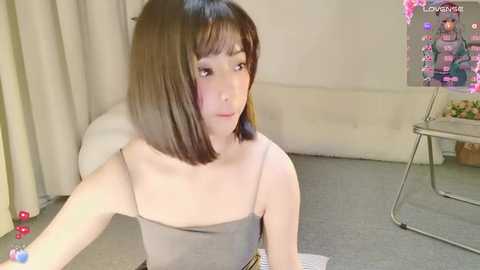 Media: A video of an East Asian woman with straight, shoulder-length brown hair, fair skin, and a slim build, wearing a gray spaghetti-strap dress, sitting on a gray carpeted floor. Background includes a white couch, a poster, and curtains.