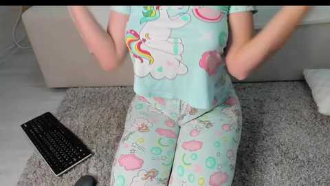 Media: Video of a person in pastel-colored unicorn pajamas, sitting on a gray carpet, hands in pockets, next to a black keyboard on a beige sofa.