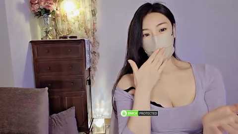 Media: Video of an East Asian woman with long black hair, wearing a light gray mask, standing in a cozy living room with a wooden dresser, flowers, and a cushioned sofa.