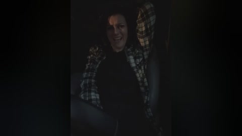 Media: A dimly lit video of a man with a medium build and fair skin, wearing a black shirt and a checkered flannel jacket, sitting in a dark car.