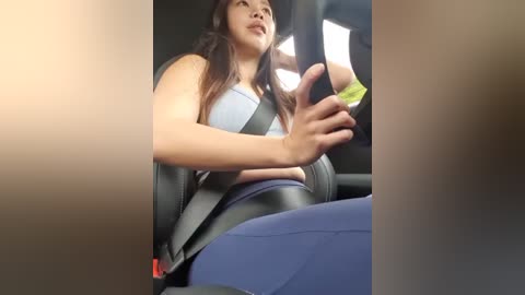 Media: Video of a young Asian woman in a car seat, wearing a light blue tank top and blue leggings, smiling, holding a green object. The car interior is dark.