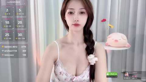 Media: Video of a young Asian woman with fair skin, wearing a lacy pink bra, braided brown hair, and a white ribbon, in a minimalist room with a pink teddy bear.