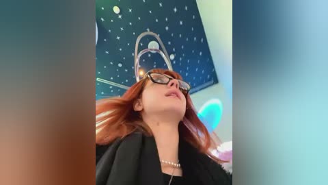 Media: Video of a young woman with long red hair, wearing black-framed glasses and a black top, gazing upward at a starry ceiling with a chandelier.