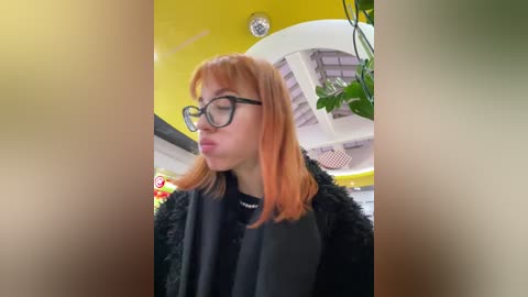 Media: Video of a young woman with fair skin, long orange hair, and large black glasses, wearing a black fur coat and black top, indoors with a yellow ceiling and green potted plant.