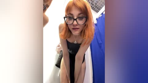 Media: Video of a slender, red-haired woman with glasses, wearing a black tank top and white towel, leaning forward in a bathroom with a white sink and blue shower curtain.