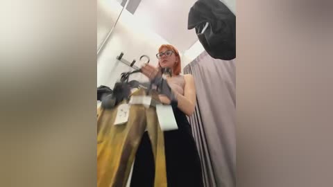 Media: A video of a red-haired woman with glasses, wearing a sleeveless black dress, standing in a changing room with a mirror, holding a pair of tan pants.