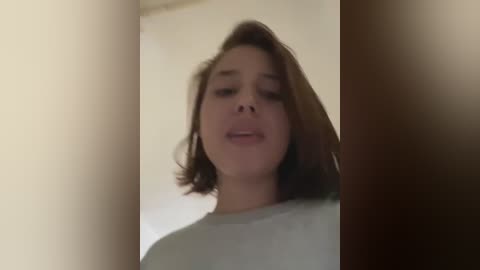 Media: A video of a young woman with light brown hair, wearing a light gray shirt, standing in a dimly lit room with beige walls. The image is slightly blurry, giving a sense of motion.