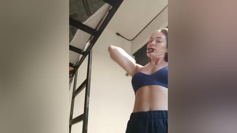 Media: Video of a fit woman with a fair complexion, wearing a dark blue sports bra and black pants, climbing a ladder in a gym with industrial ceiling beams.