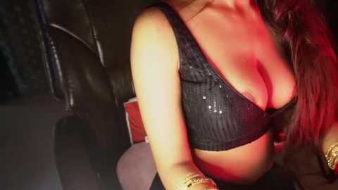 Media: Video of a woman with long, wavy red hair, wearing a black, sequined bra that highlights her ample cleavage, sitting in a dimly lit room with red lighting.