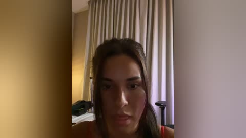 Media: Video of a young woman with straight, dark brown hair, light skin, and a neutral expression. She is indoors, likely in a hotel room, with beige curtains and a bed in the background.