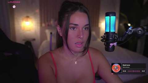 Media: A video of a young woman with fair skin, dark hair, and red spaghetti strap top, using a blue-lit microphone in a dimly-lit bedroom.