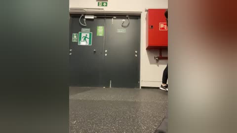 Media: Video of a dimly lit, utilitarian hallway with gray doors, green emergency signs, a red fire extinguisher, and a partially visible person in dark clothing in the background.