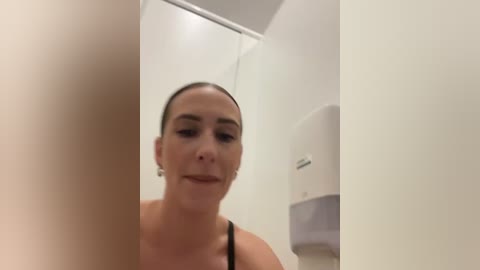 Media: Video of a woman with light skin and brown hair in a ponytail, wearing a black tank top, looking at a mirror in a white-tiled bathroom with a paper towel dispenser.