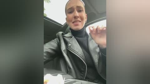 Media: Video of a light-skinned woman with short hair, wearing a black leather jacket and turtleneck, seated in a car. She is smiling and holding a cigarette. The background shows a blurry outdoor scene.