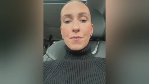 Media: A video of a bald, light-skinned woman with a neutral expression, wearing a black turtleneck sweater, sitting in a car. The background features a blurred interior with grey upholstery.