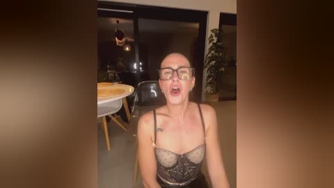 Media: Video of a man with a shaved head, wearing glasses and a black lace bra, sitting in a chair, with a messy face and open mouth, in a dimly lit room with a round table and black chairs.