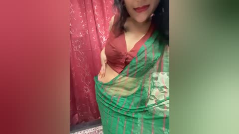 Media: Video of a woman with medium skin tone, wearing a green and red saree, exposing a red bra, standing against a red and green curtain.