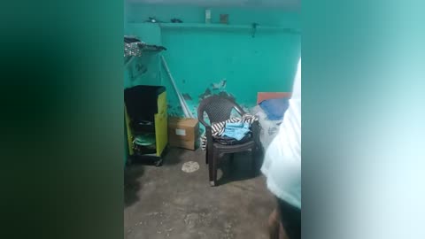 Media: Video of a small, dimly lit room with a turquoise wall. The floor is bare concrete. Various items including a black chair with a blue shirt, a yellow trolley, a zebra-striped pillow, and a transparent umbrella are scattered about.