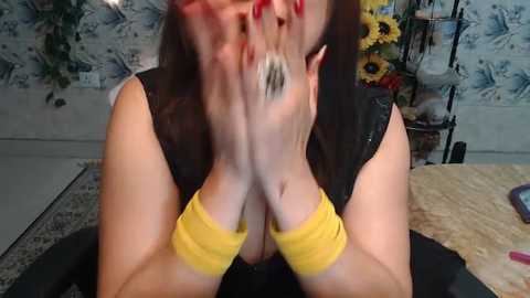 Media: A video captures a woman with long brown hair, wearing a black sleeveless top, covering her face with her hands. She has yellow bracelets and red-painted nails. Background includes blue floral wallpaper, sunflowers, and a beige rug.