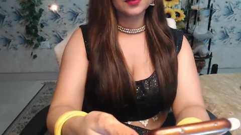 Media: Video of a woman with long brown hair, wearing a black sequin top, yellow bracelets, and a silver necklace, seated in a room with floral wallpaper and a sunflower arrangement.