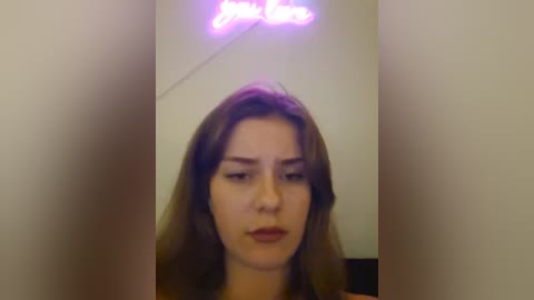 Media: Video of a young woman with fair skin and straight brown hair, looking contemplative with slight pout, against a plain white background. A neon sign reading \"I Love You\" hangs above her head.