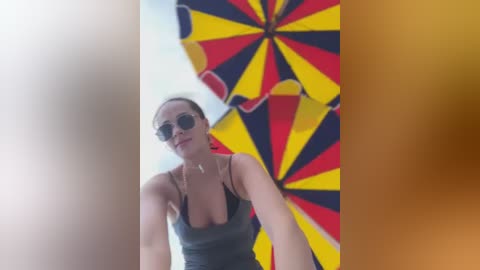 Media: A video features a woman with a slender physique, wearing a grey tank top and aviator sunglasses, holding a large, colorful umbrella with yellow, red, and black stripes. The background is blurred.