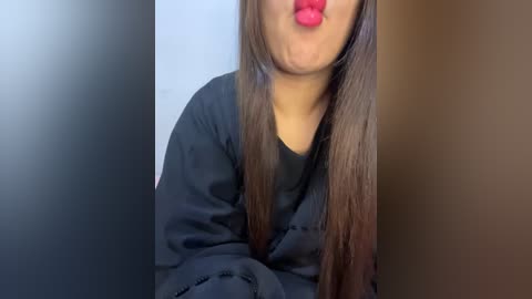 Media: Video of a young woman with long, straight brown hair, wearing a black, long-sleeved shirt, puckering her lips with bright red lipstick. The background is a plain, light-colored wall.