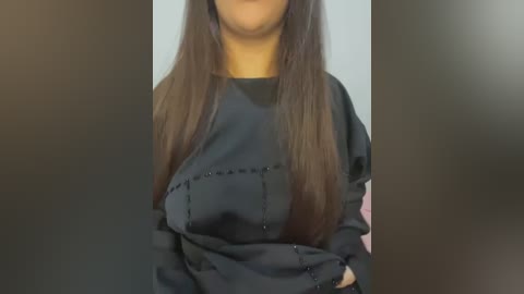 Media: Video of a woman with long, straight brown hair, wearing a black, long-sleeved shirt, with a hint of a pink garment visible on the right side. Background is blurred.