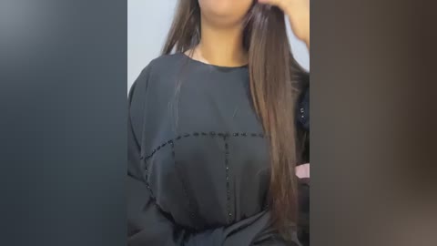 Media: A video featuring a woman with medium skin tone and long, straight brown hair. She wears a black, long-sleeved top with intricate, vertical stitching and a subtle, feminine pattern. The background is blurred, focusing on her upper body and torso.