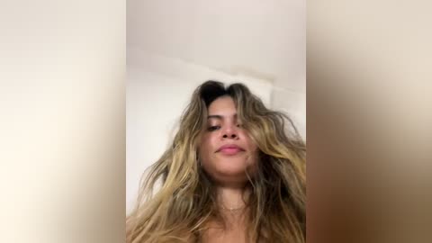 Media: Video of a young woman with long, wavy, light brown hair, standing in a minimalist, white-walled room. She has a neutral expression and is wearing a soft, light-colored top.