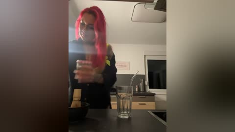 Media: A video of a woman with long, bright pink hair, wearing a black jacket, taking a selfie in a modern kitchen. The background features white walls, a window, and a stainless steel stove.